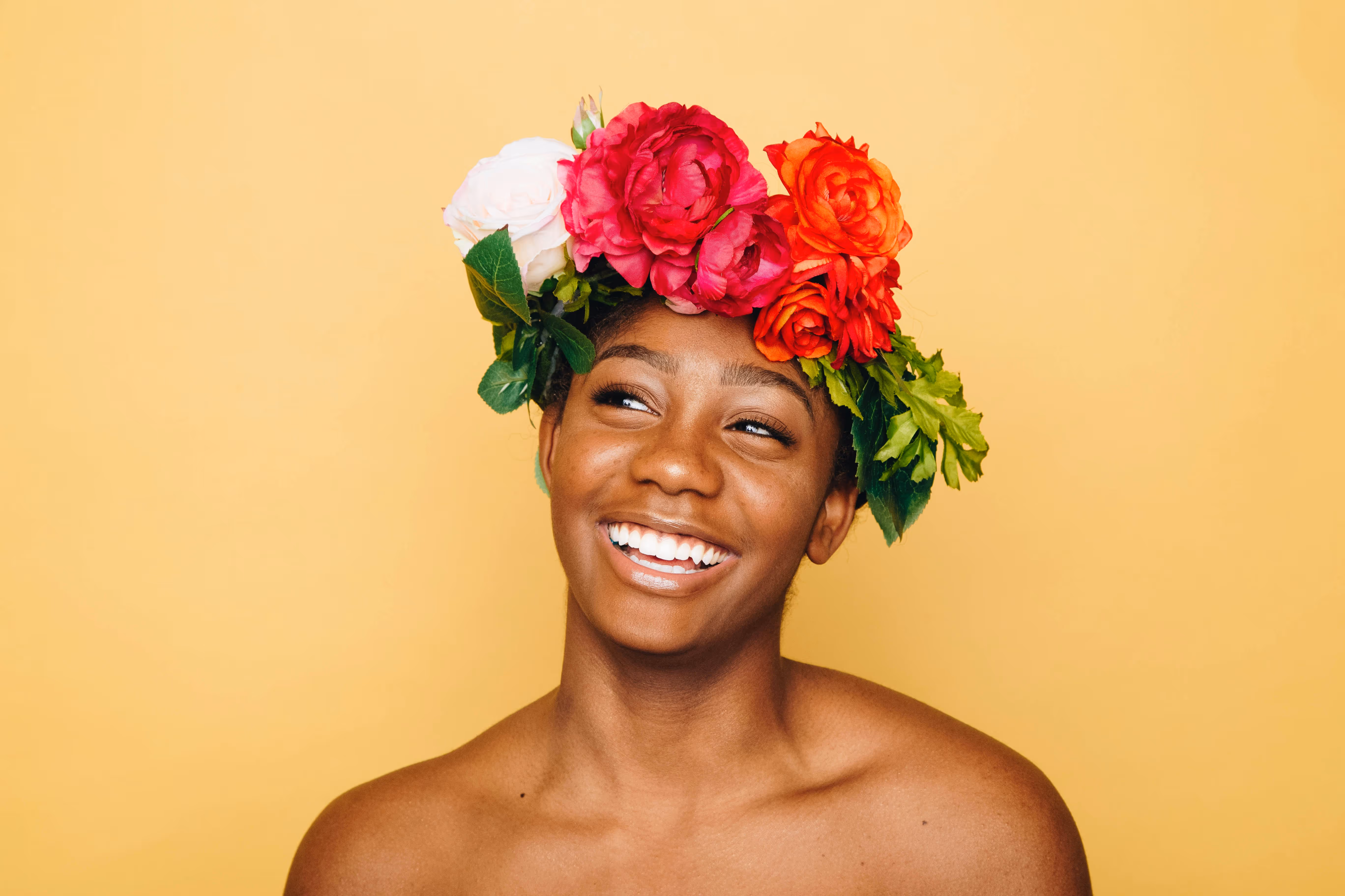 Smiling woman with dark skin wearing a colorful flower crown against a yellow background.