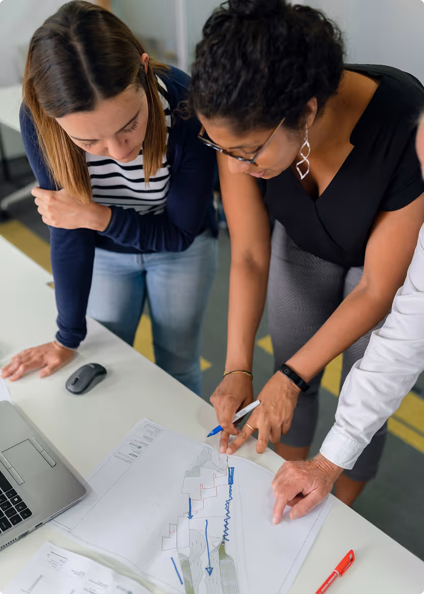 Three colleagues closely examining and discussing a blueprint on a desk with a laptop and stationery.