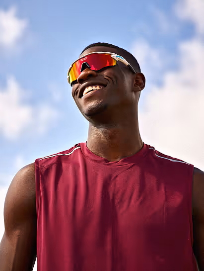 Smiling young athlete wearing red sports sunglasses and a sleeveless maroon shirt outdoors with a blue sky background.