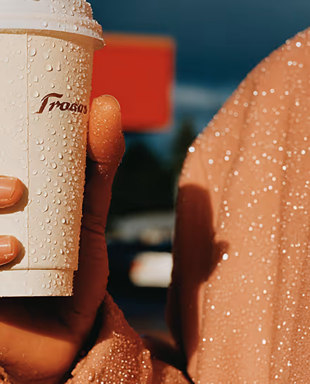 Hand holding a white takeaway coffee cup covered in raindrops, with a rain-drenched sleeve visible against a blurred dark background.