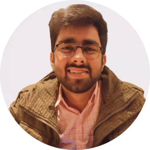 Smiling man with glasses, beard, and mustache wearing a brown jacket and pink checkered shirt.