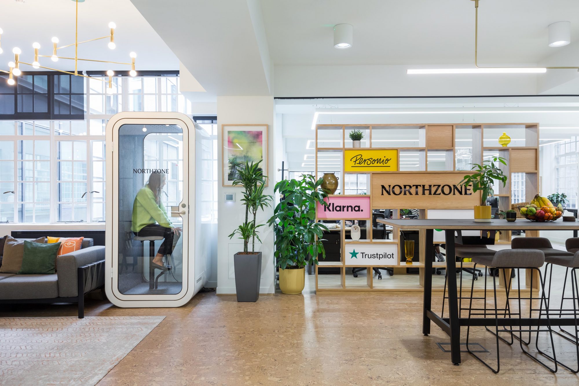 Modern office space with a woman sitting inside a transparent phone booth labeled Northzone, a gray sofa, plants, and a wooden shelf displaying logos of Personio, Klarna, and Trustpilot.