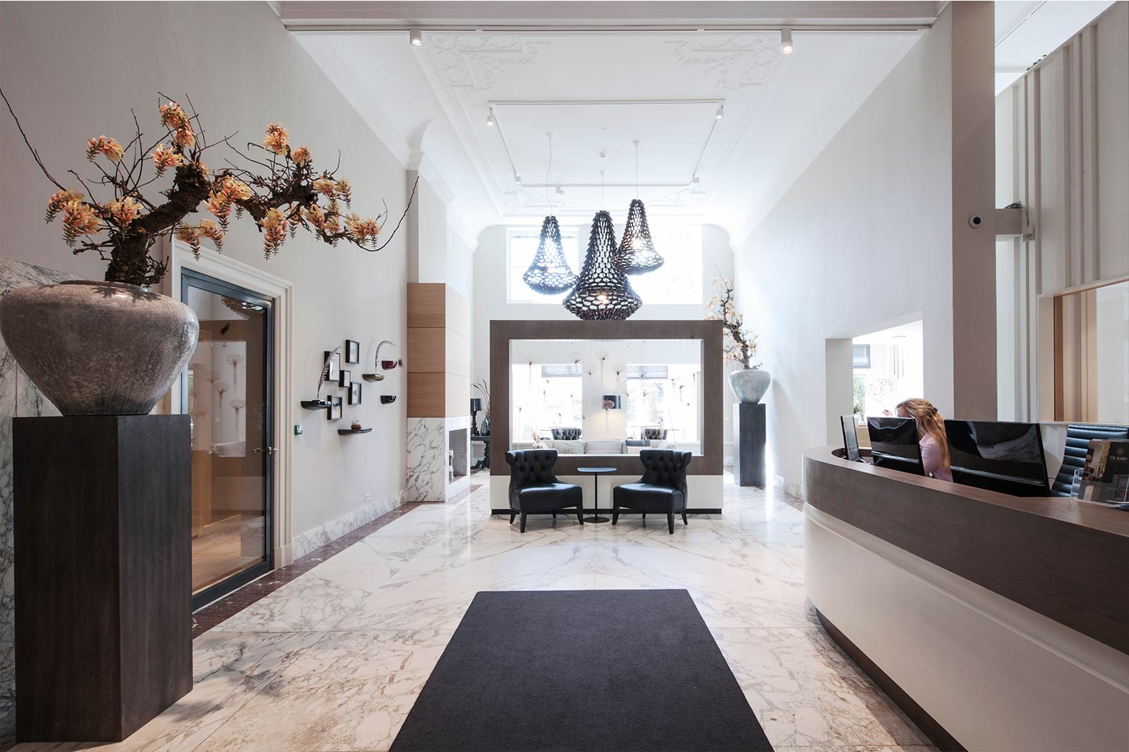 Bright hotel lobby with marble floors, a reception desk, black chairs, modern pendant lights, and decorative flower arrangements.