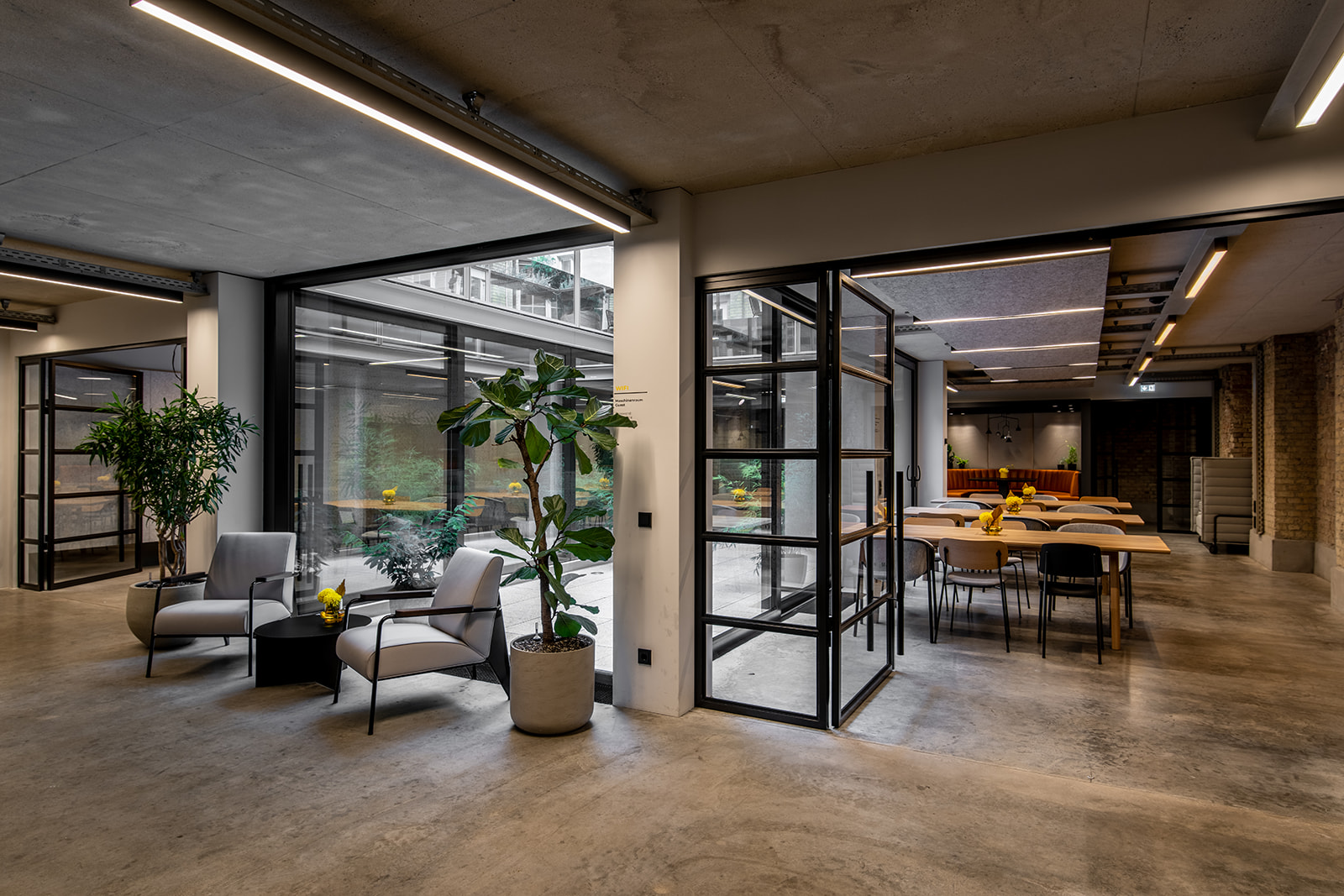 Modern office lounge area with gray armchairs, potted plants, and a meeting room with wooden tables and chairs visible through glass walls.