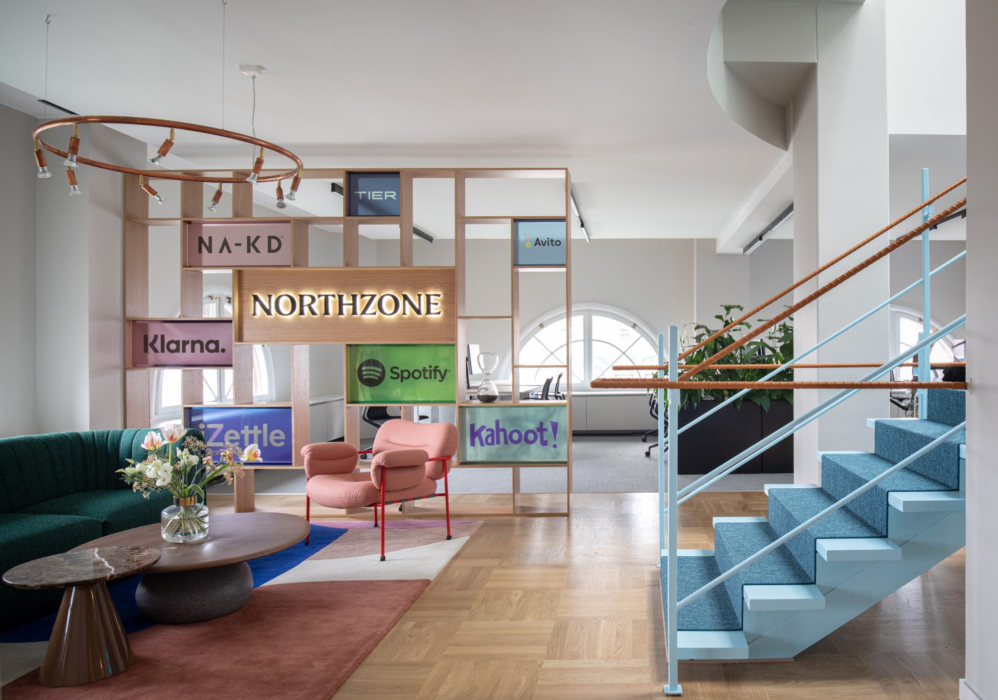 Modern office lounge with a green curved sofa, pink armchair, colorful rug, wooden partition displaying logos of companies including Northzone, Spotify, and Klarna, and blue-carpeted staircase.