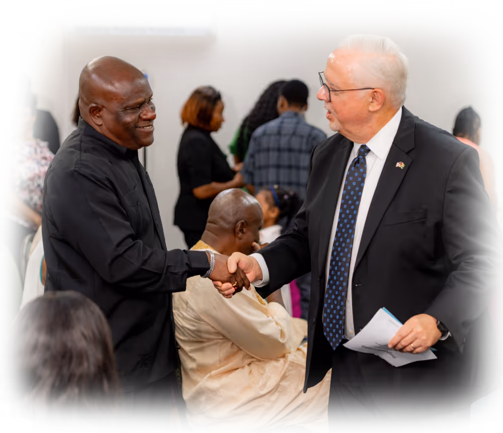Two men shaking hands in a room with seated and standing people in the background, one man wearing a black shirt and the other in a suit with a blue patterned tie.