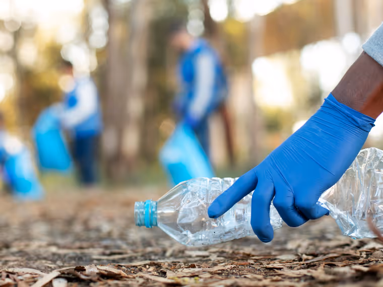 Person wearing a blue glove picking up a plastic bottle litter in a forest with others collecting trash in the background.