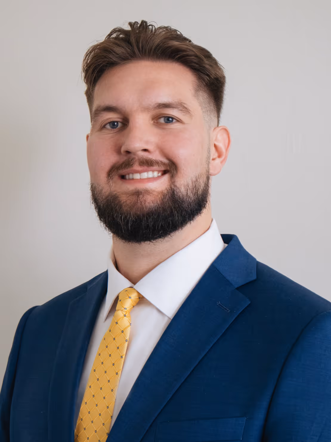 Smiling man with beard wearing a blue suit jacket, white shirt, and yellow patterned tie against a gray background.