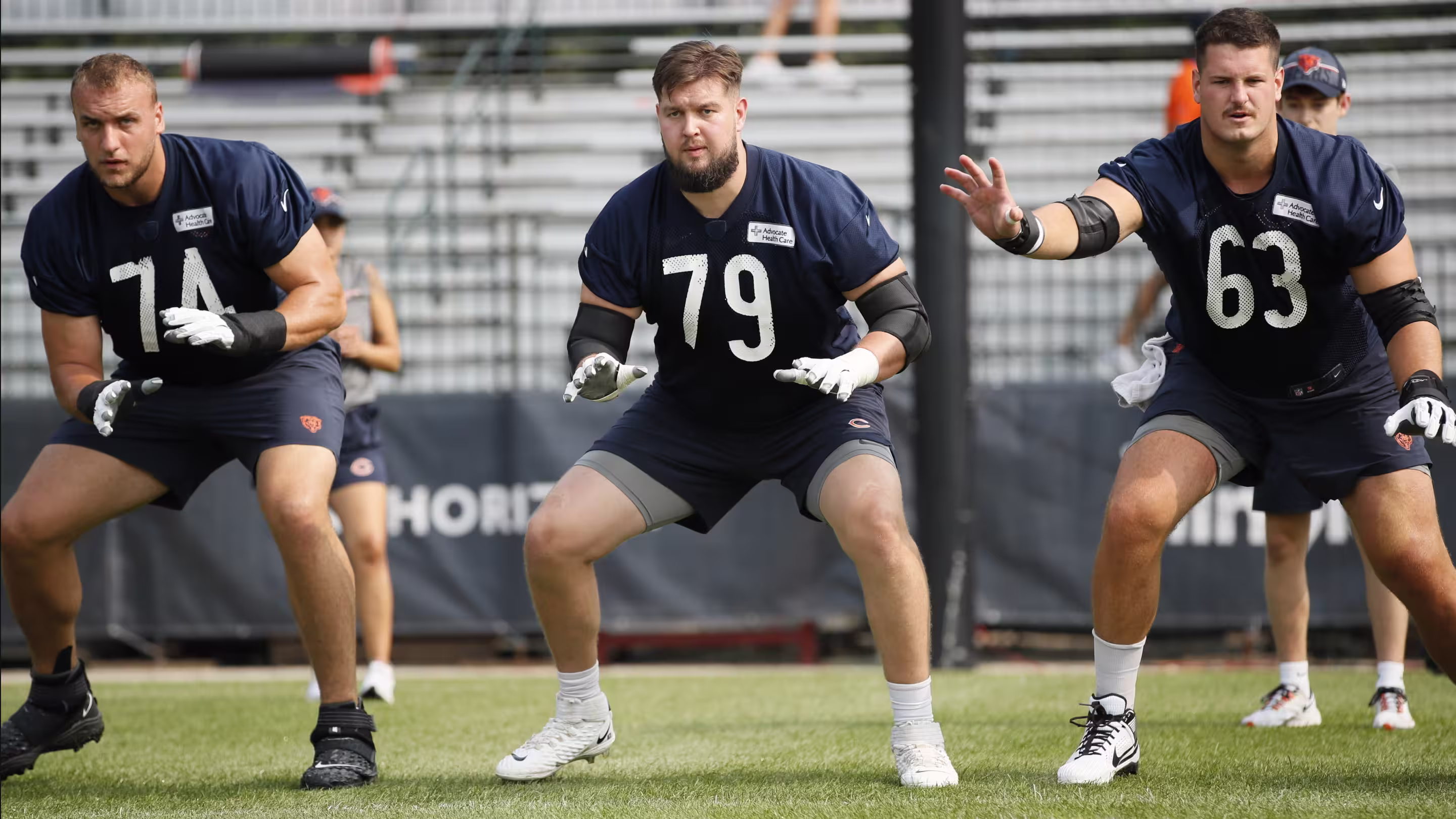 Three male football players in navy practice uniforms in stance on a field during training.