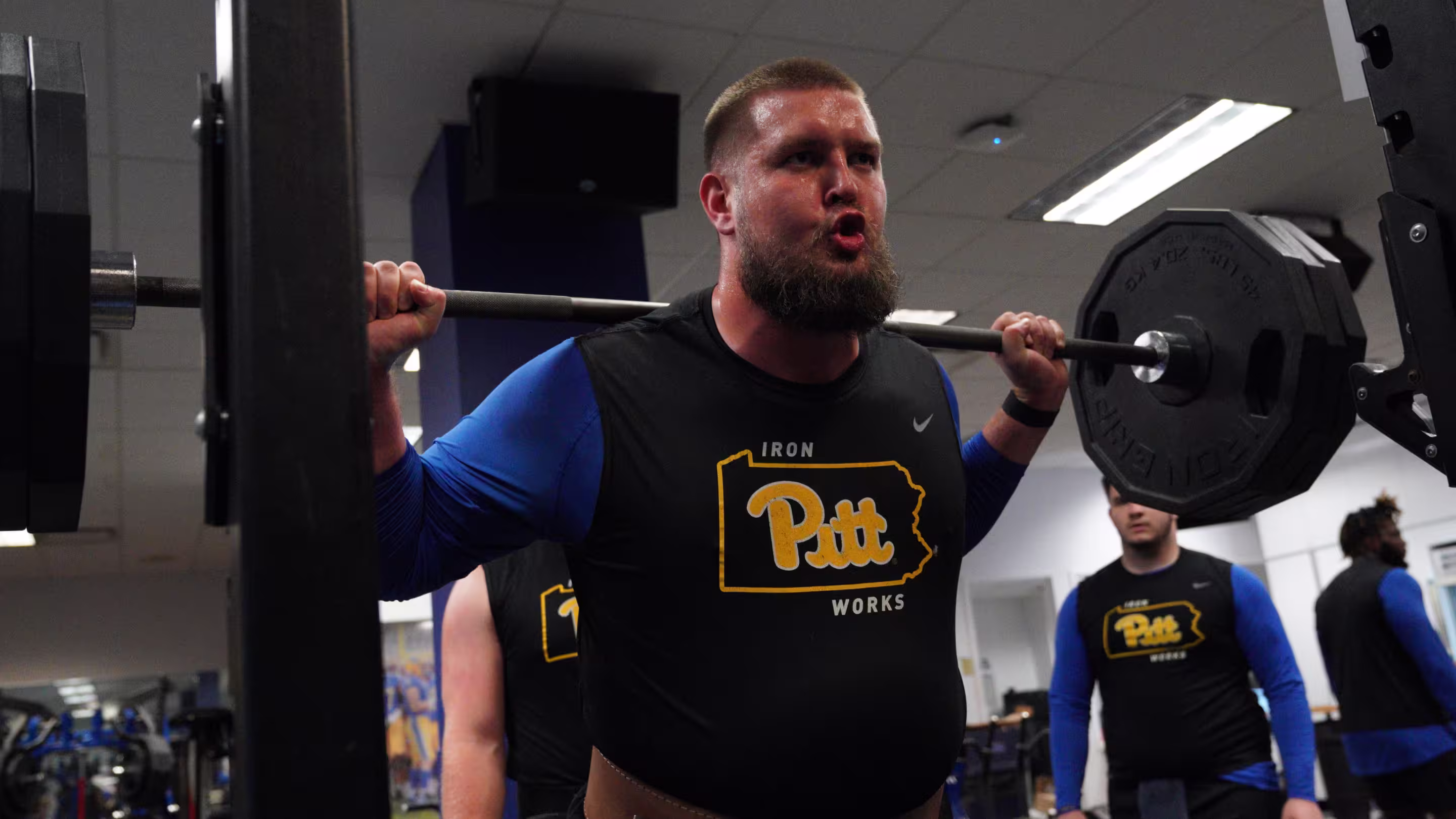 Bearded man lifting a heavily weighted barbell on his shoulders in a gym while wearing a black Pitt Iron Works shirt.