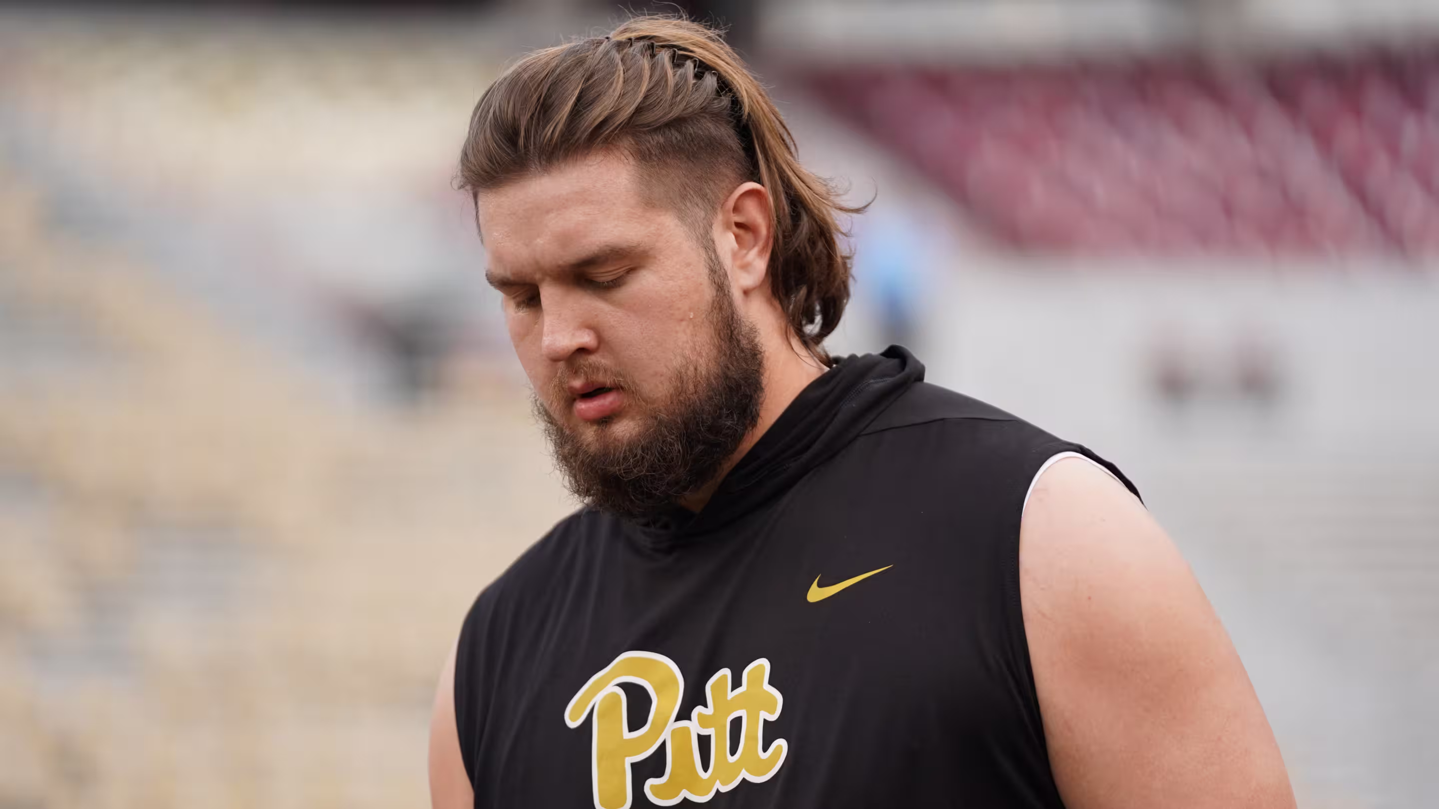 Man with braided hair and beard wearing a black sleeveless hoodie with 'Pitt' and a gold Nike logo.