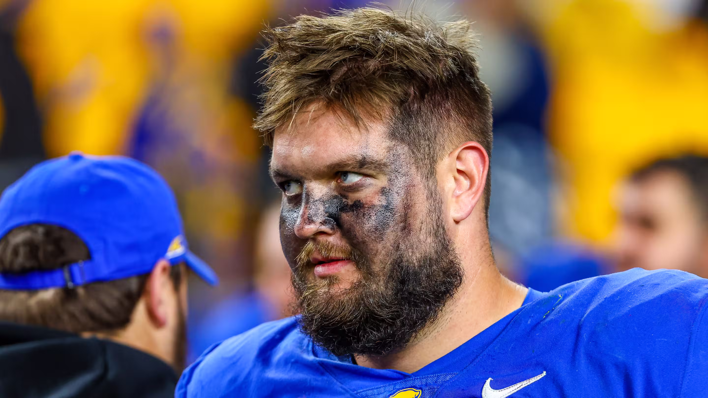 Close-up of a male football player with black eye paint and a beard, wearing a blue jersey during a game.