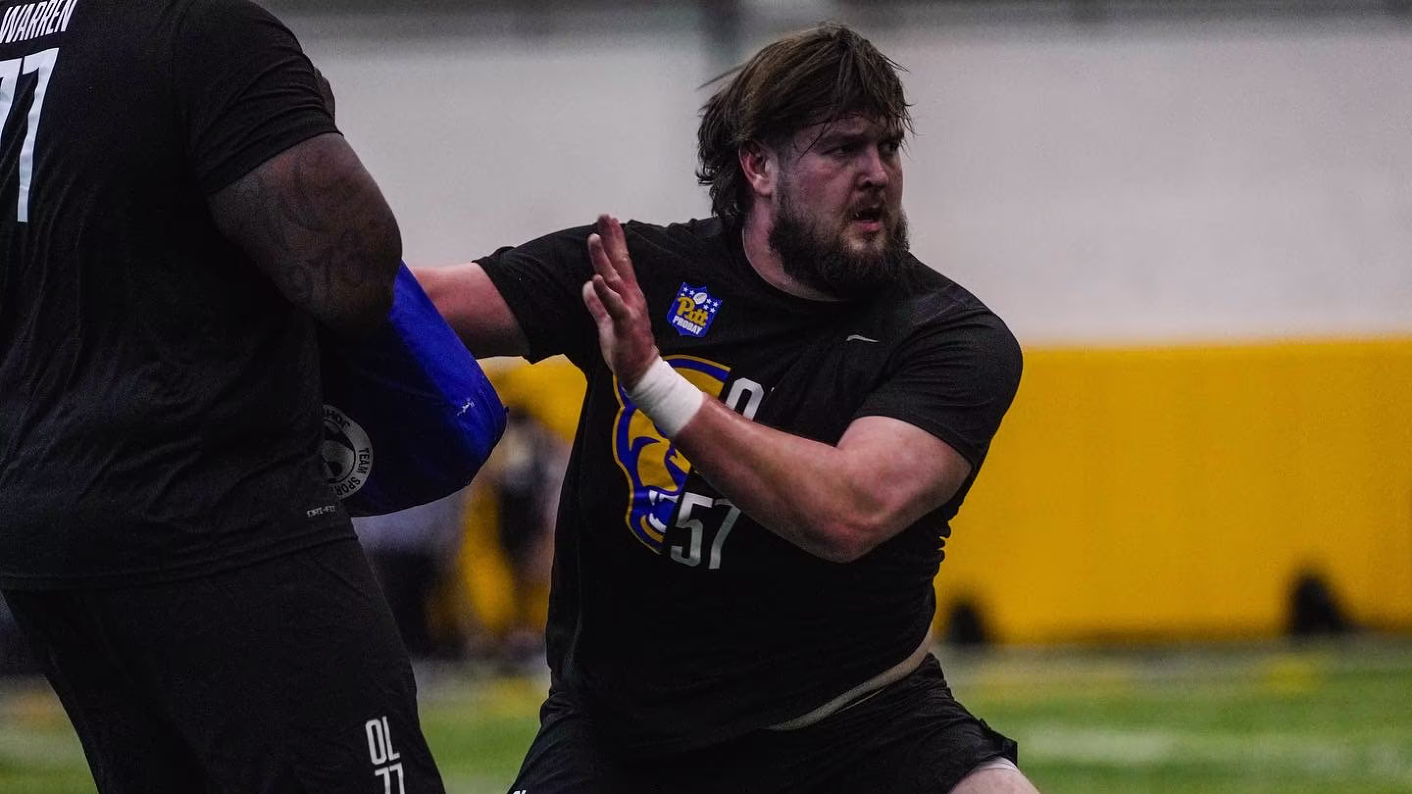 Football player in black training gear pushing against a blue blocking pad held by another player during indoor practice.