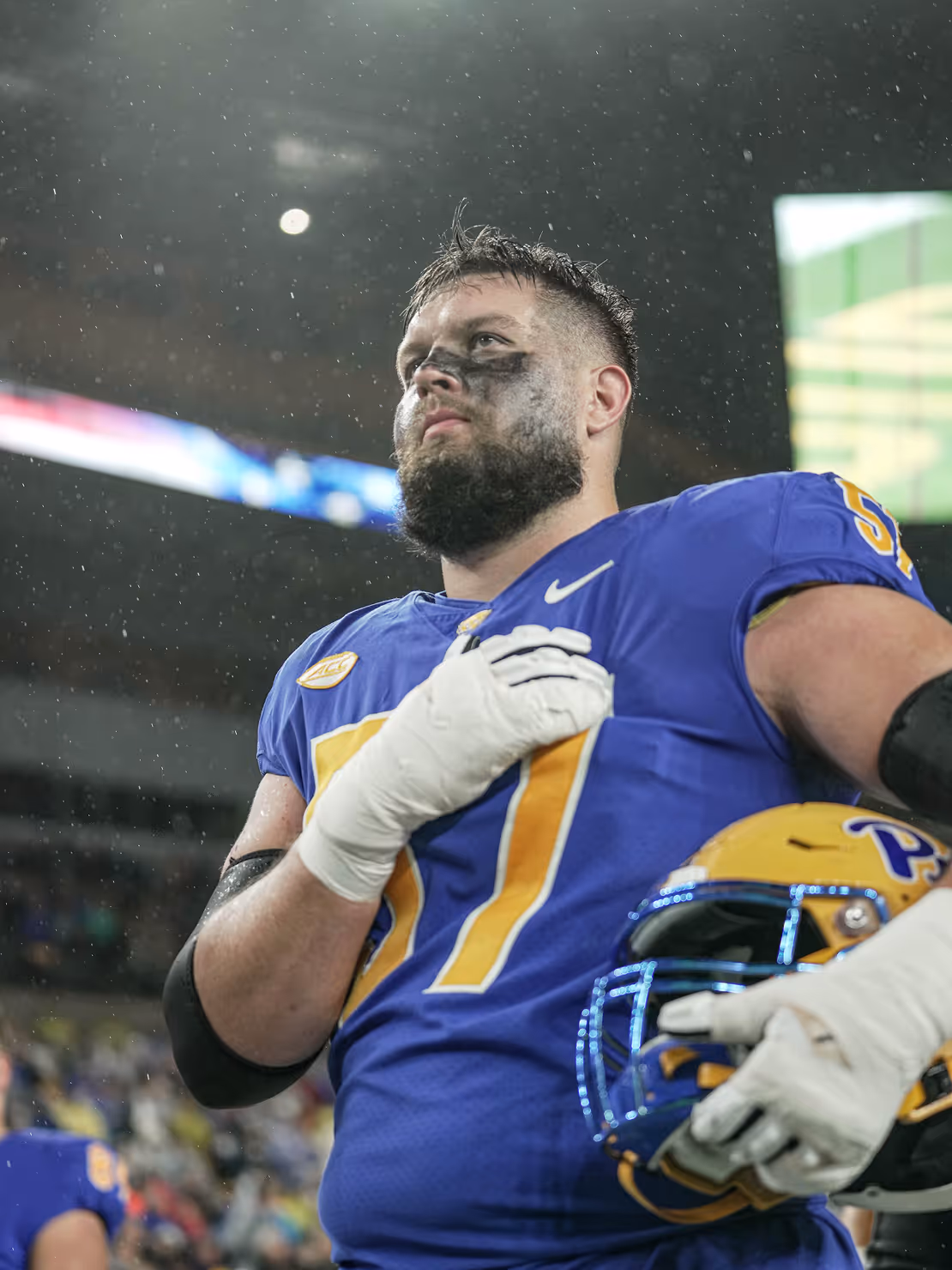 Football player in blue and yellow uniform with black eye paint holds helmet and places gloved hand over chest.