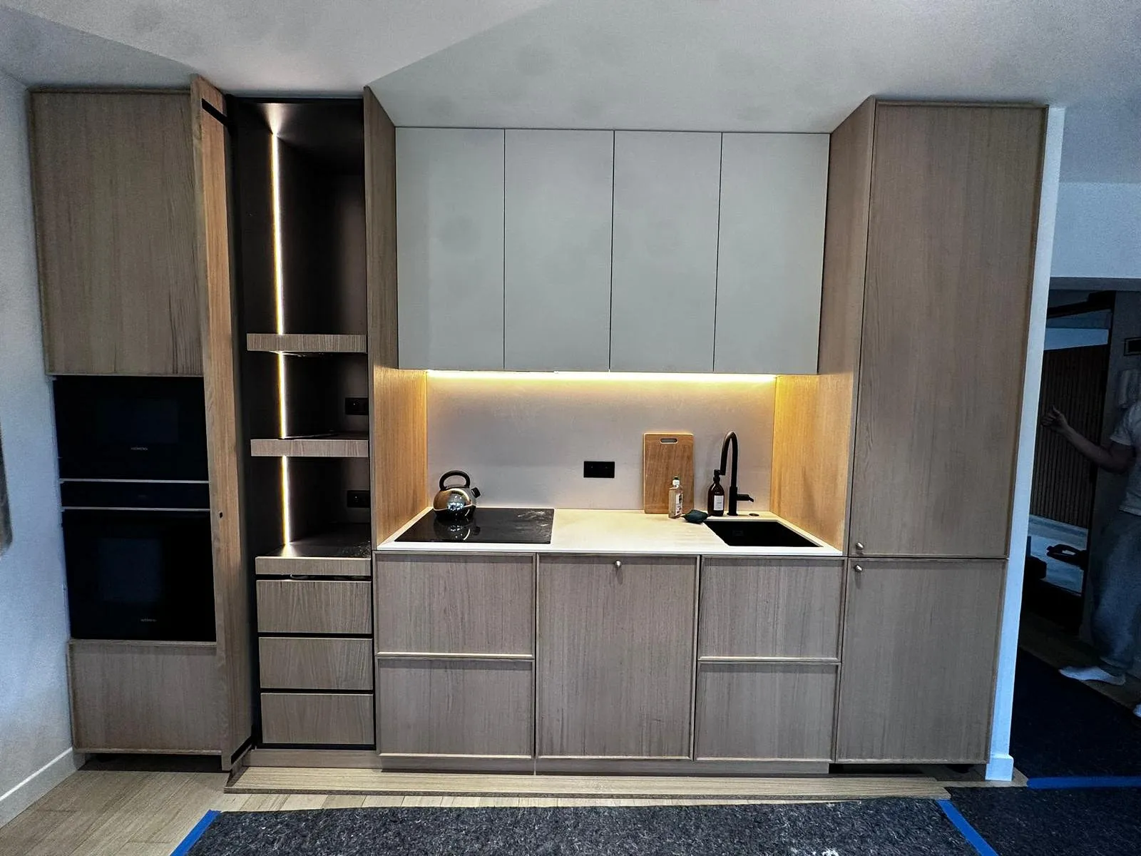 Cuisine moderne avec armoires en bois clair, plaque de cuisson noire, évier noir, théière métallique et éclairage LED sous les étagères.