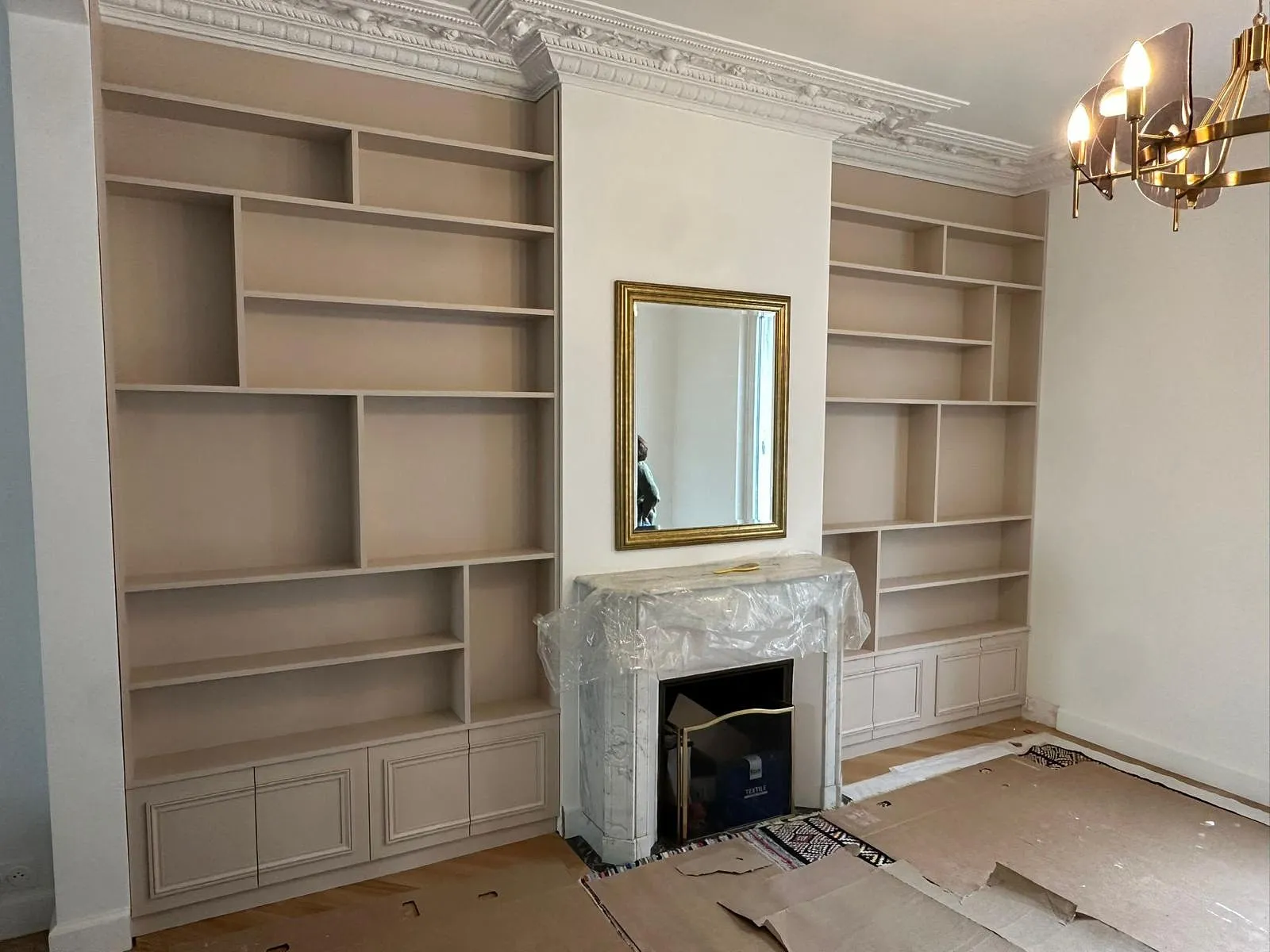 Pièce avec deux grandes étagères encadrant une cheminée en marbre blanche, miroir doré au-dessus, et sol protégé par du carton.