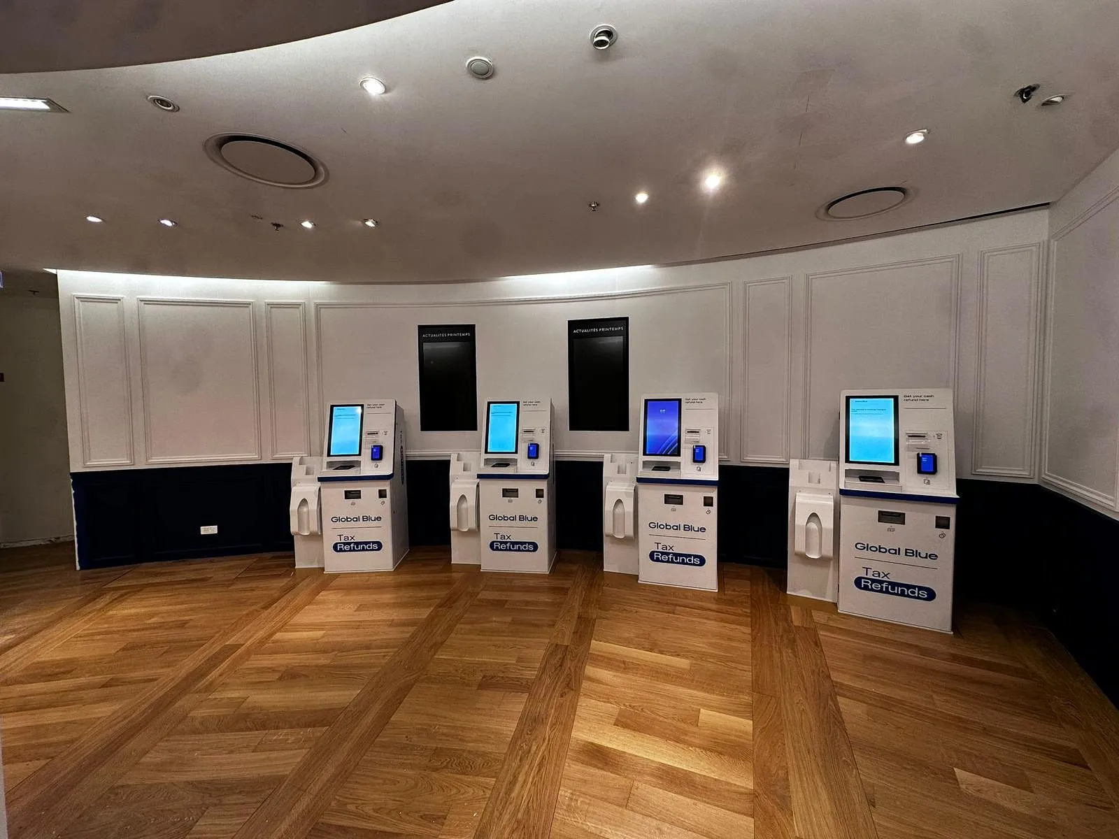 Quatre bornes blanches Global Blue pour remboursements de taxes alignées sur un sol en parquet, mur blanc et éclairage au plafond.