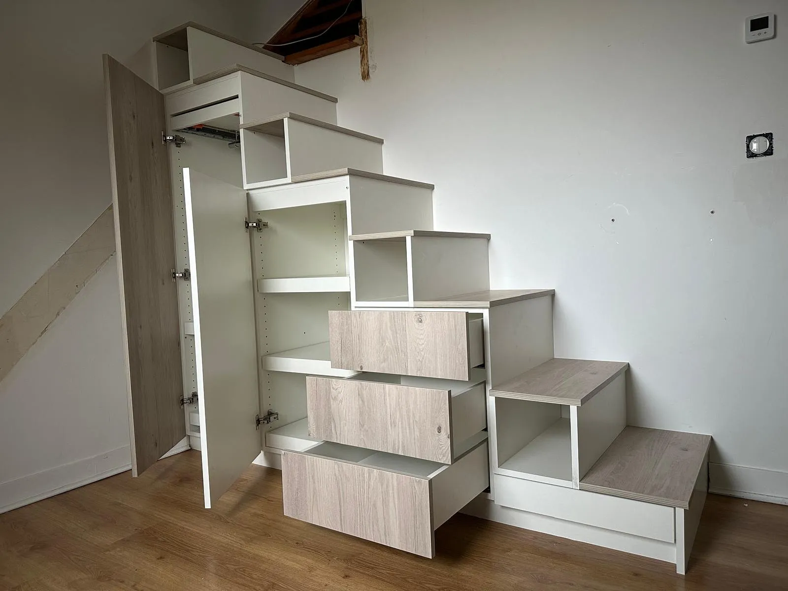 Escalier compact en bois clair avec rangements intégrés comprenant des tiroirs ouverts et une porte de placard.
