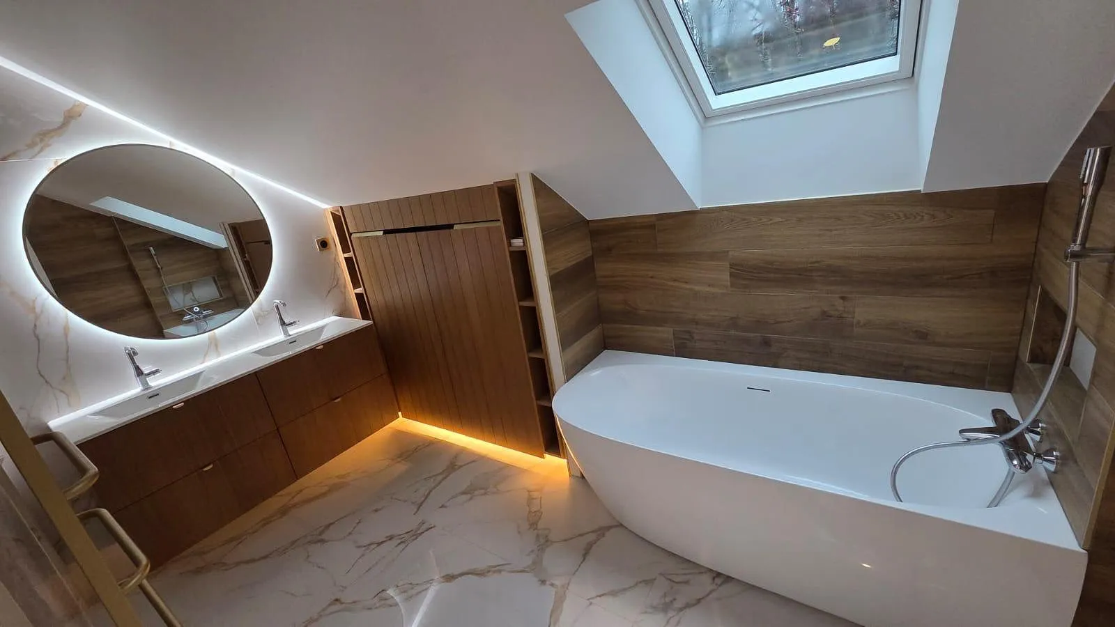 Salle de bain moderne avec baignoire blanche autonome, grand miroir rond rétroéclairé, double lavabo sur meuble en bois et fenêtre de plafond.