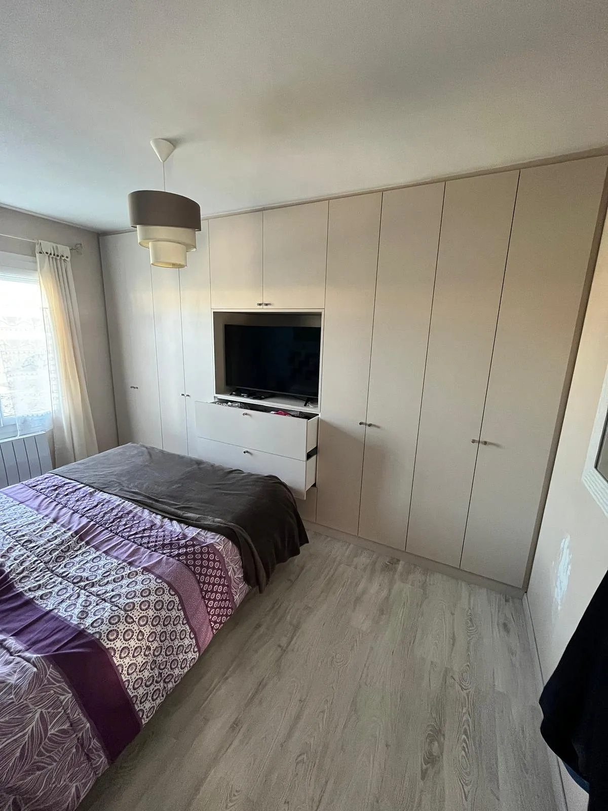Chambre avec lit couvert d'une couette violette et beige, un grand placard beige avec un téléviseur encastré et deux tiroirs ouverts, sol en parquet clair, et fenêtre avec rideaux blancs.