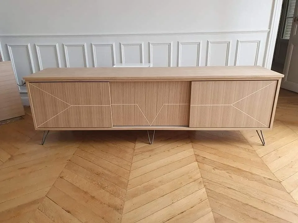 Meuble bas en bois clair avec 3 portes à motifs géométriques, sur un sol en parquet chevron.
