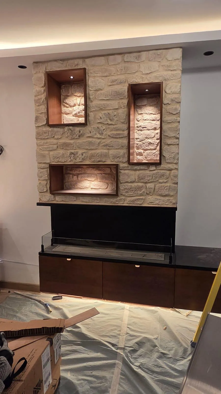 Mur en pierre avec trois niches éclairées encastrées au-dessus d'une cheminée moderne noire avec rangement bois en bas, sol protégé par une bâche.