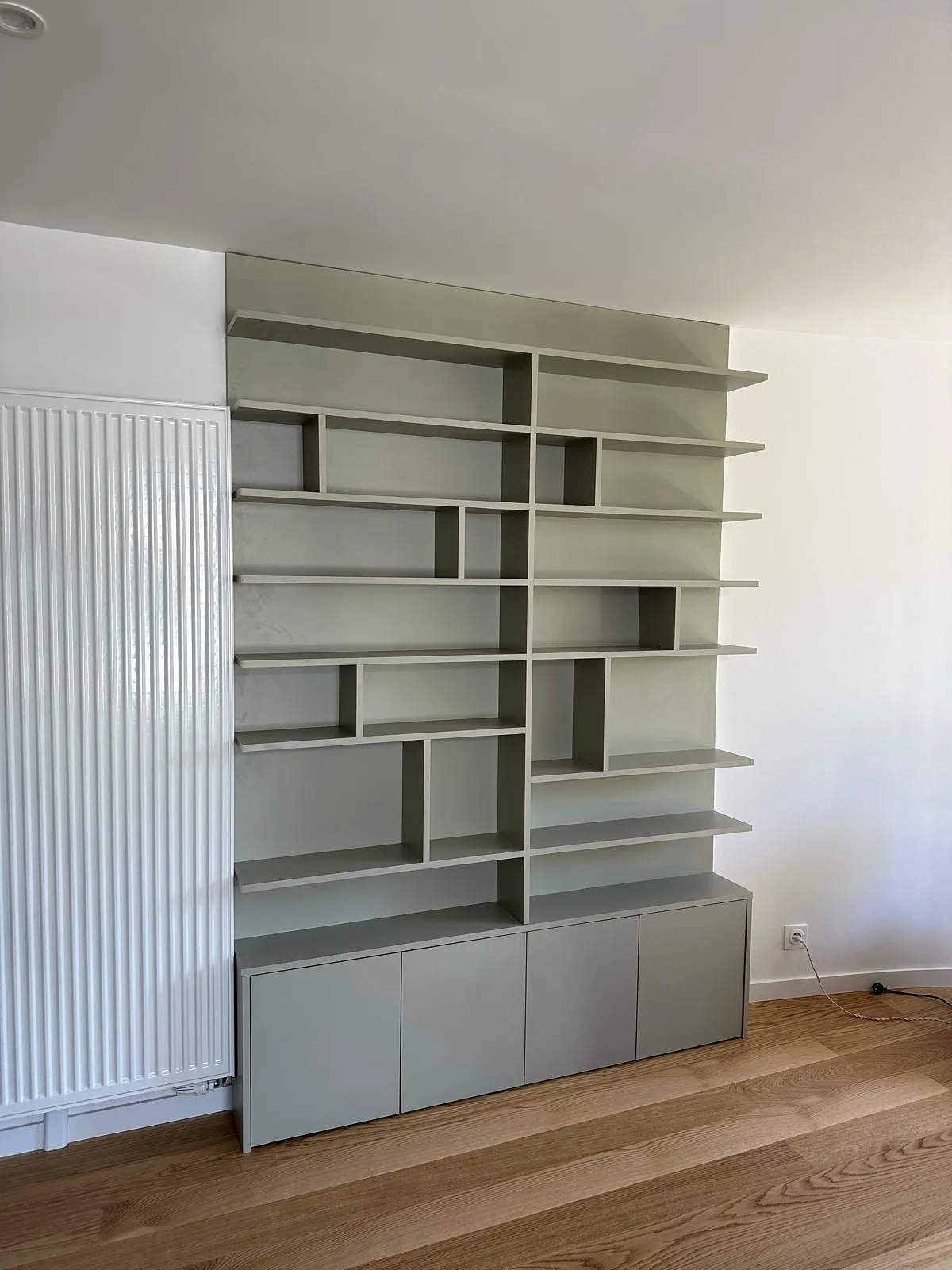 Étagère murale grise moderne avec multiples compartiments ouverts et placards en bas, posée sur un sol en bois clair à côté d'un radiateur blanc.