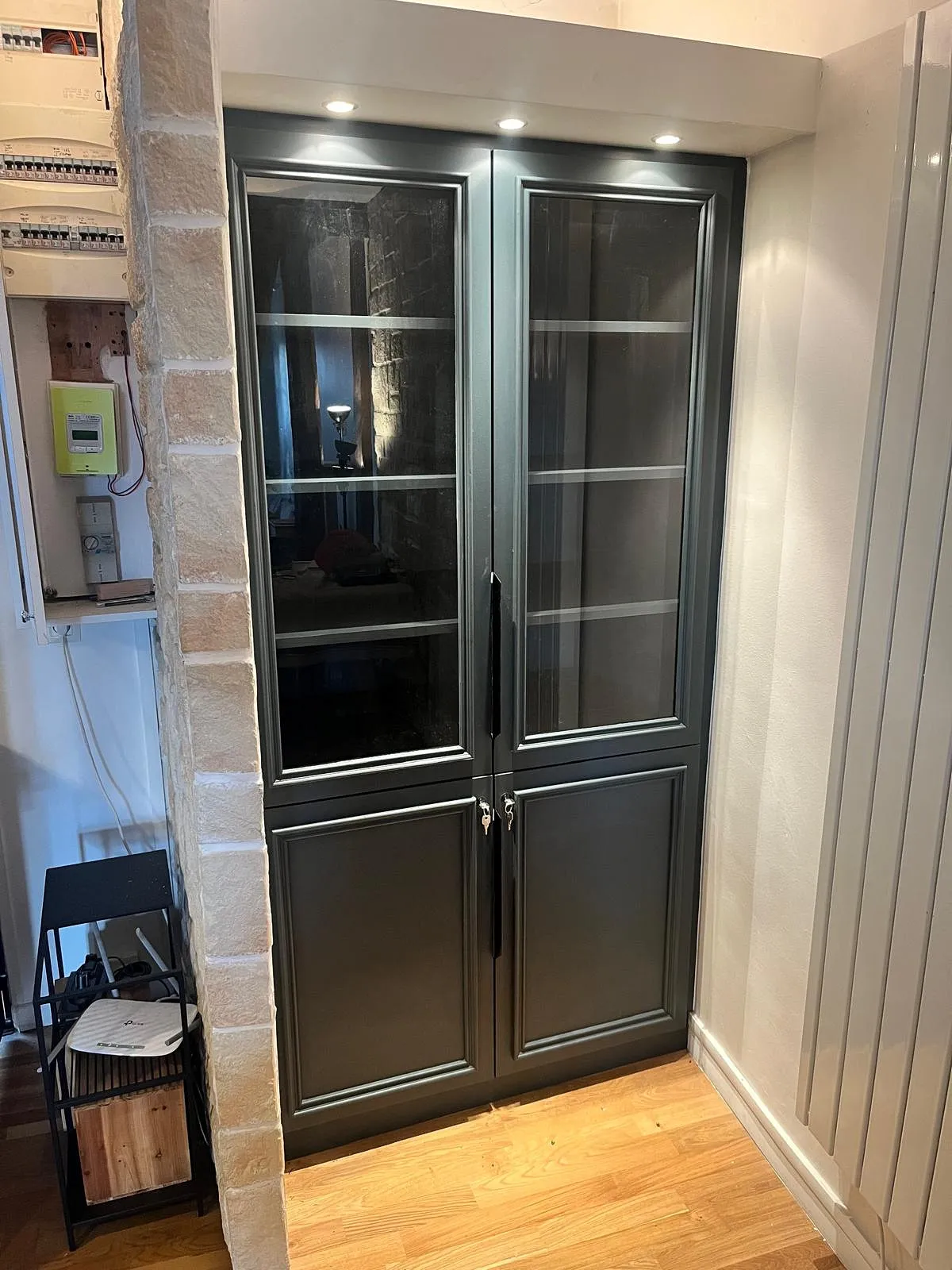 Placard gris avec portes vitrées sous un luminaire encastré dans un coin avec mur en pierre et sol en parquet.