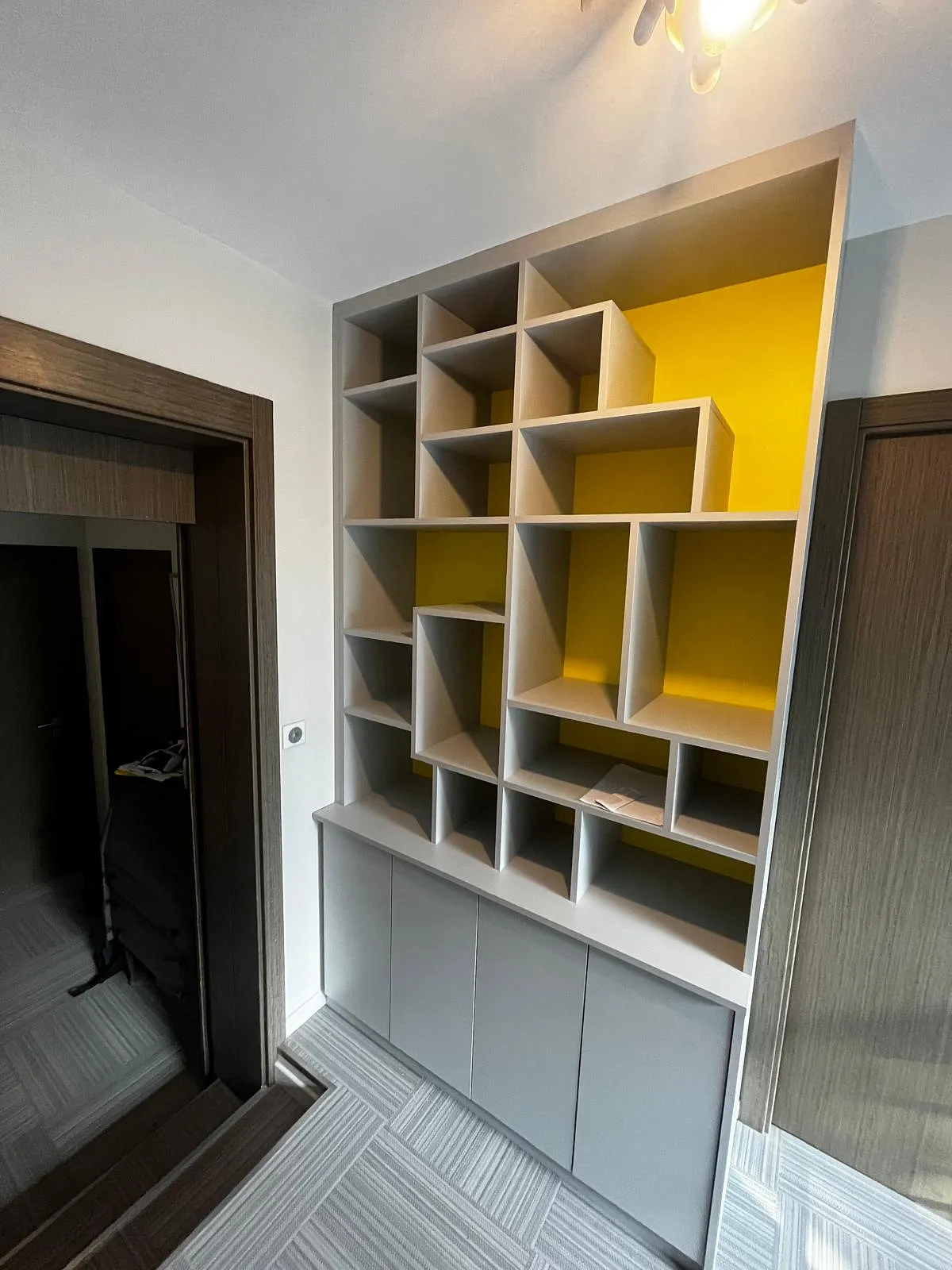 Étagère grise avec compartiments de formes variées et un mur jaune en arrière-plan, entre deux portes en bois foncé dans un couloir.