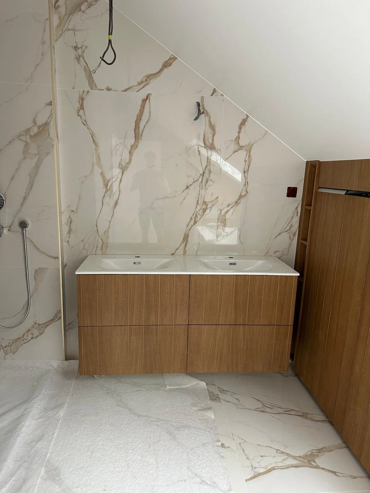 Salle de bain avec un double lavabo moderne sur meuble en bois, murs et sol en carrelage blanc marbré, et douche à main sur la gauche.