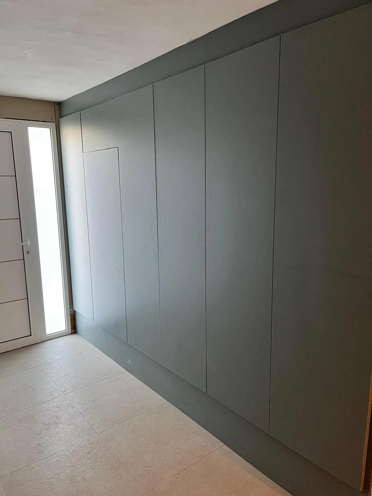 Un mur avec plusieurs panneaux gris uniformes et une porte blanche avec fenêtre latérale, sol en carrelage clair.