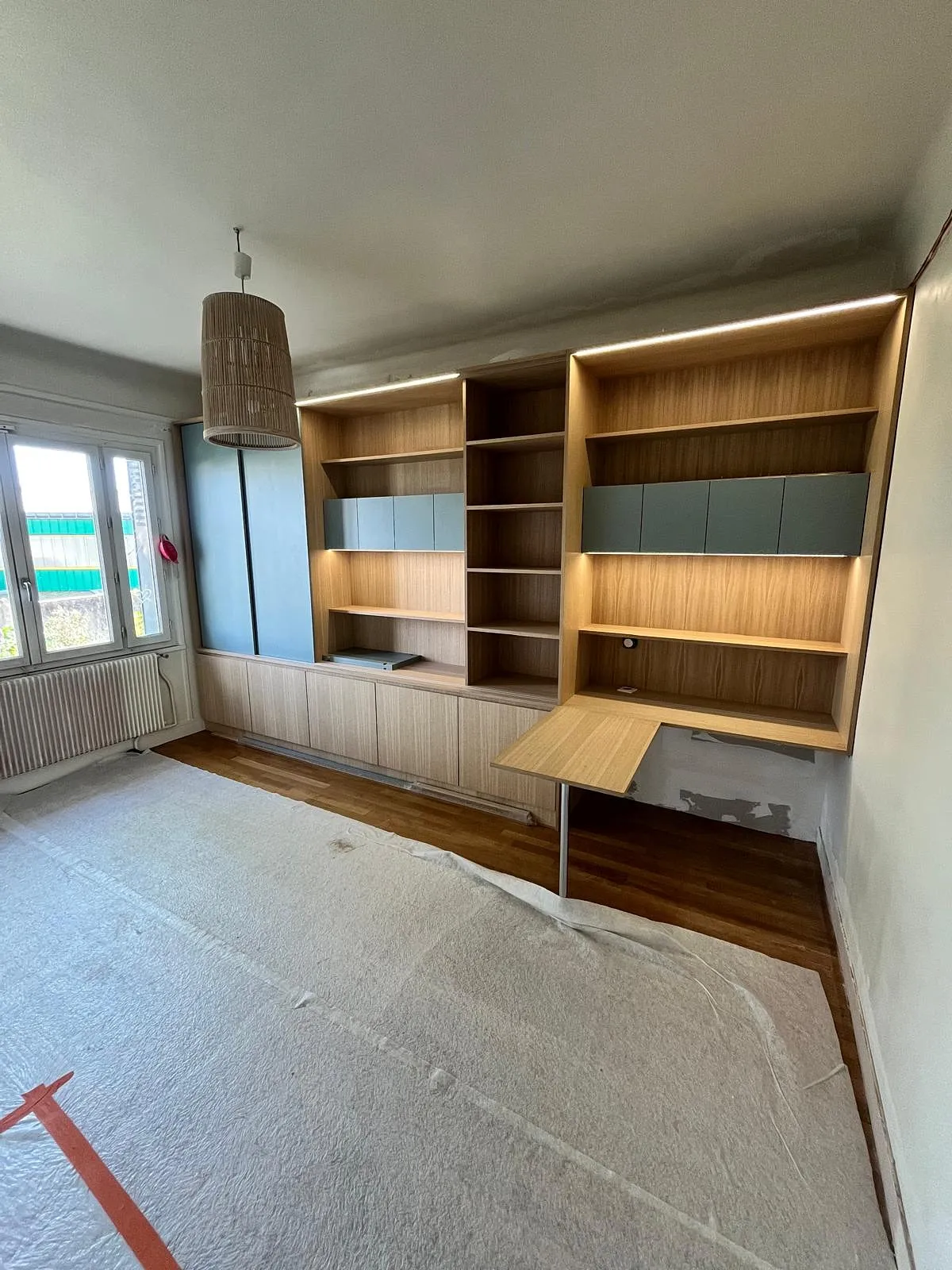 Pièce avec grand meuble mural en bois clair comprenant étagères, armoires et un bureau pliant, sol protégé par une couverture, fenêtre à gauche.