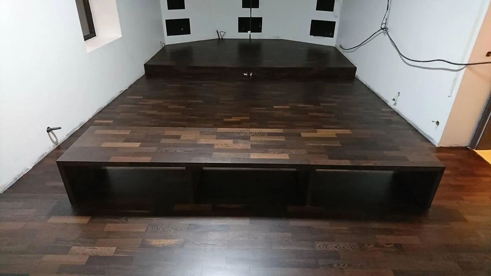 Une estrade en bois sombre avec trois compartiments ouverts en dessous dans une pièce aux murs blancs.