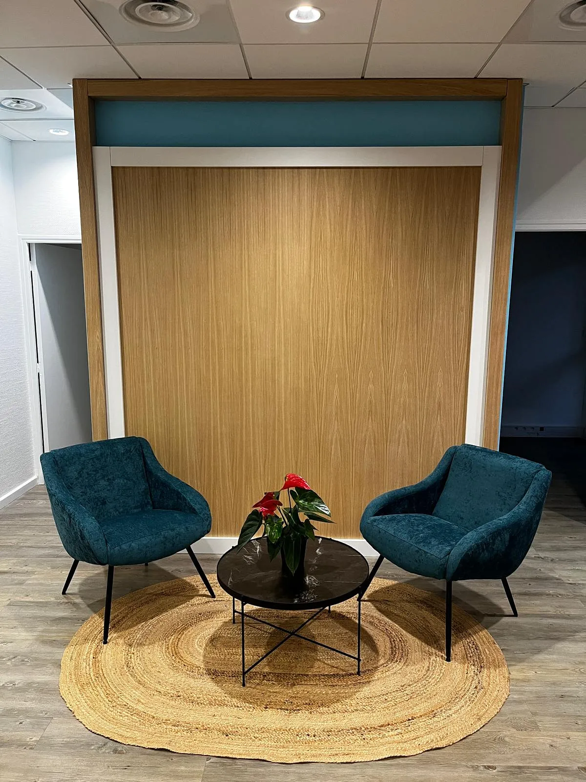 Deux fauteuils bleus foncés encadrent une table basse ronde noire avec plante à fleurs rouges, sur un tapis rond en jute, devant un panneau mural en bois clair.