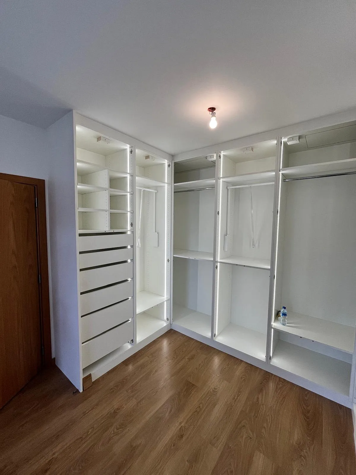 Dressing moderne avec étagères et tiroirs blancs intégrés, sol en bois et porte en bois à gauche.