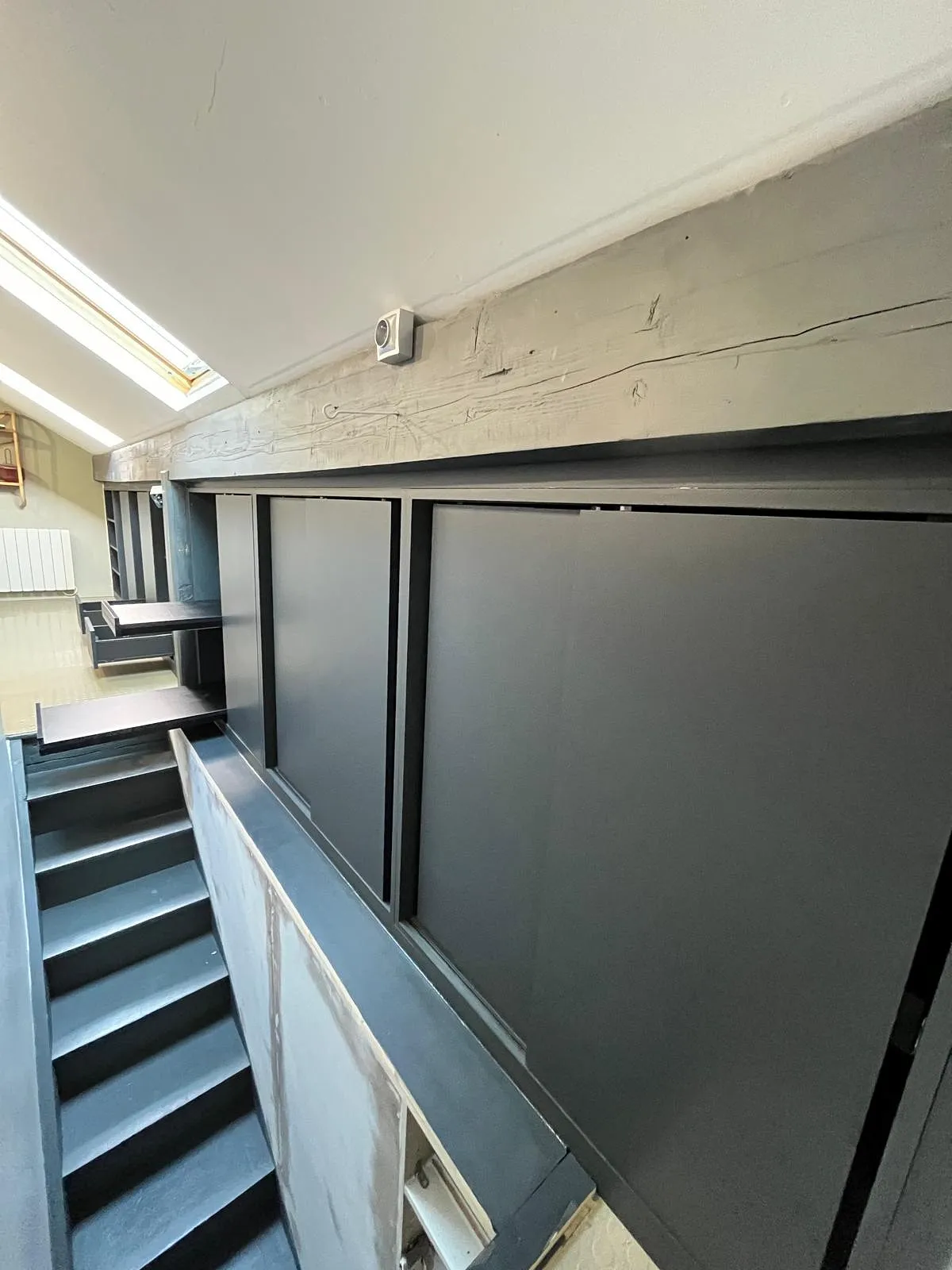 Escalier intérieur étroit avec murs et panneaux gris foncé sous un plafond incliné avec une fenêtre de toit.