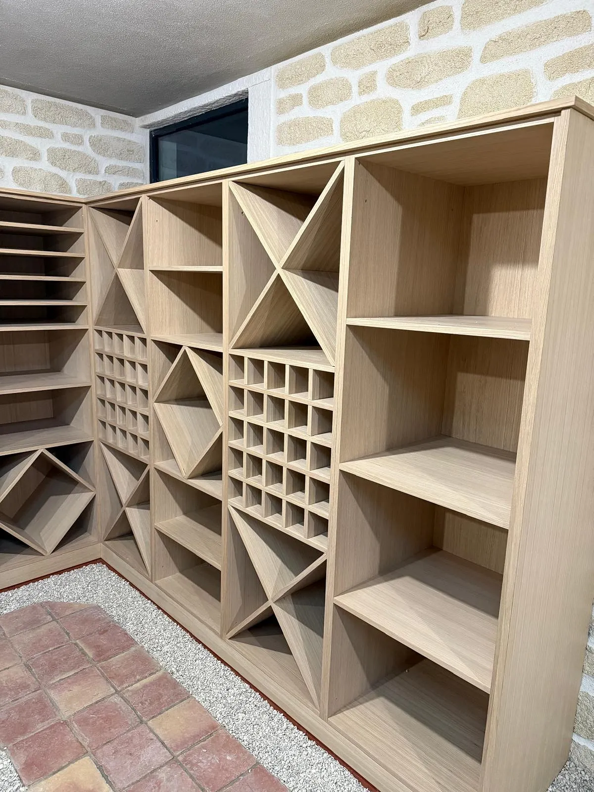 Étagère en bois avec multiples compartiments de formes variées dans une pièce avec mur en pierre blanche.