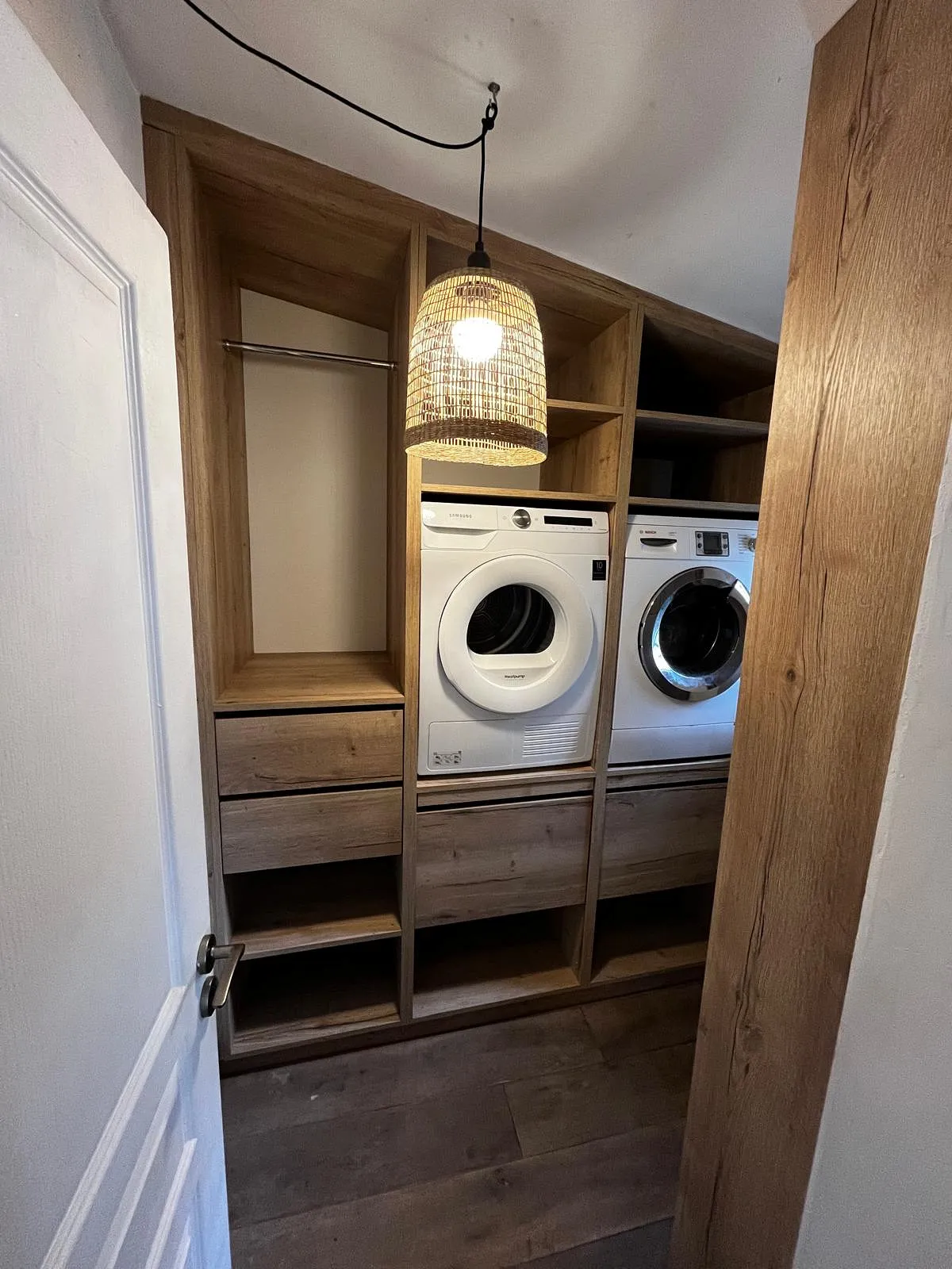 Buanderie avec lave-linge et sèche-linge blancs encastrés dans des étagères en bois, lampadaire suspendu en osier, sol en bois.