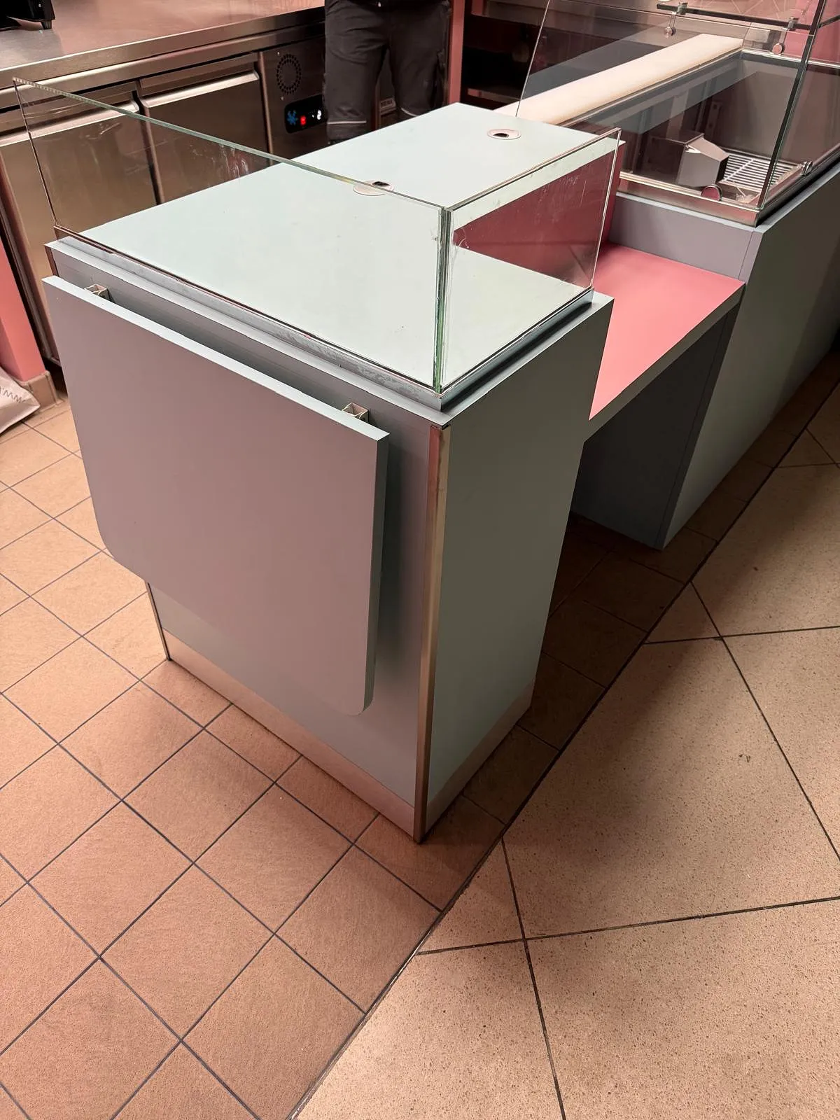 Comptoir en métal gris avec vitrine en verre sur un sol carrelé, partie d'un espace commercial ou de restauration.