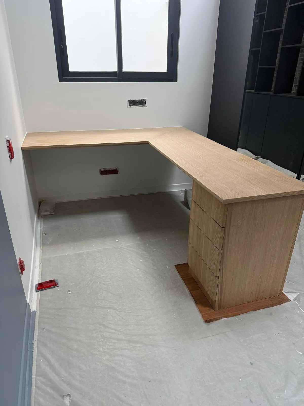 Bureau en angle en bois clair installé dans une pièce avec une fenêtre noire, sol protégé par une bâche plastique.