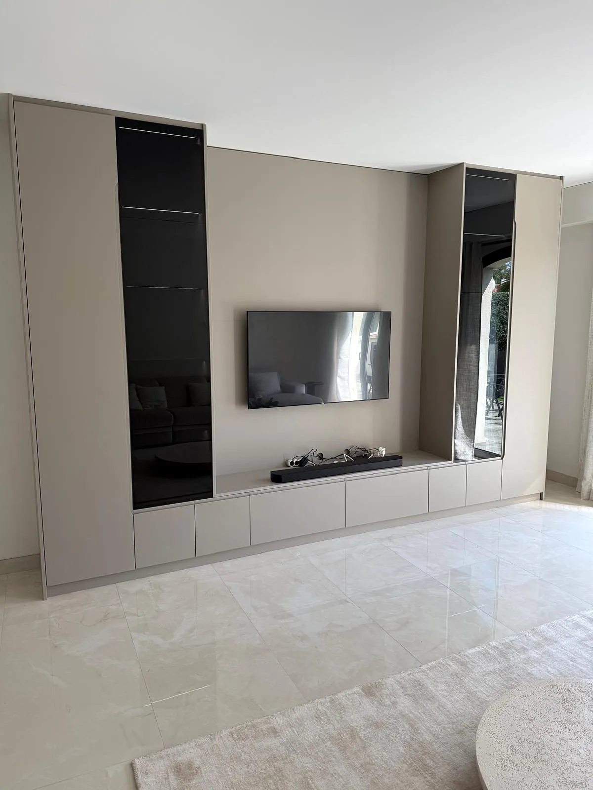 Meuble TV moderne beige avec deux colonnes vitrées noires, TV murale, barre de son, sol en carrelage brillant et tapis clair.
