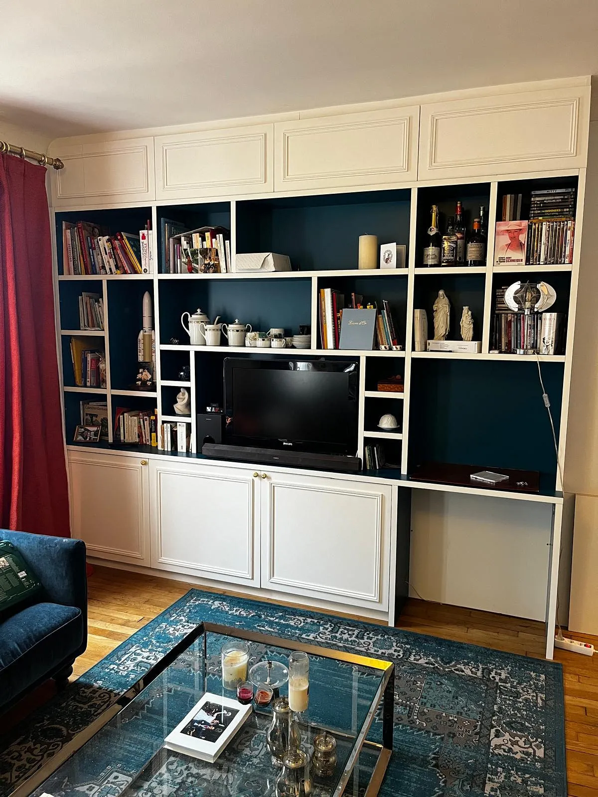 Bibliothèque blanche avec étagères ouvertes contenant des livres, une télé, des objets de décoration, un service à thé et des bouteilles, à côté d'un canapé bleu et d'une table basse en verre avec des bougies et des livres.