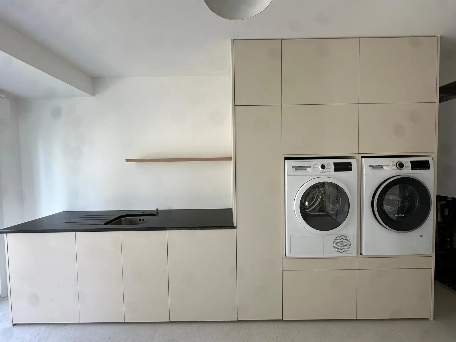 Buanderie moderne avec lave-linge et sèche-linge encastrés dans des meubles beige, avec un plan de travail noir et un évier intégré.
