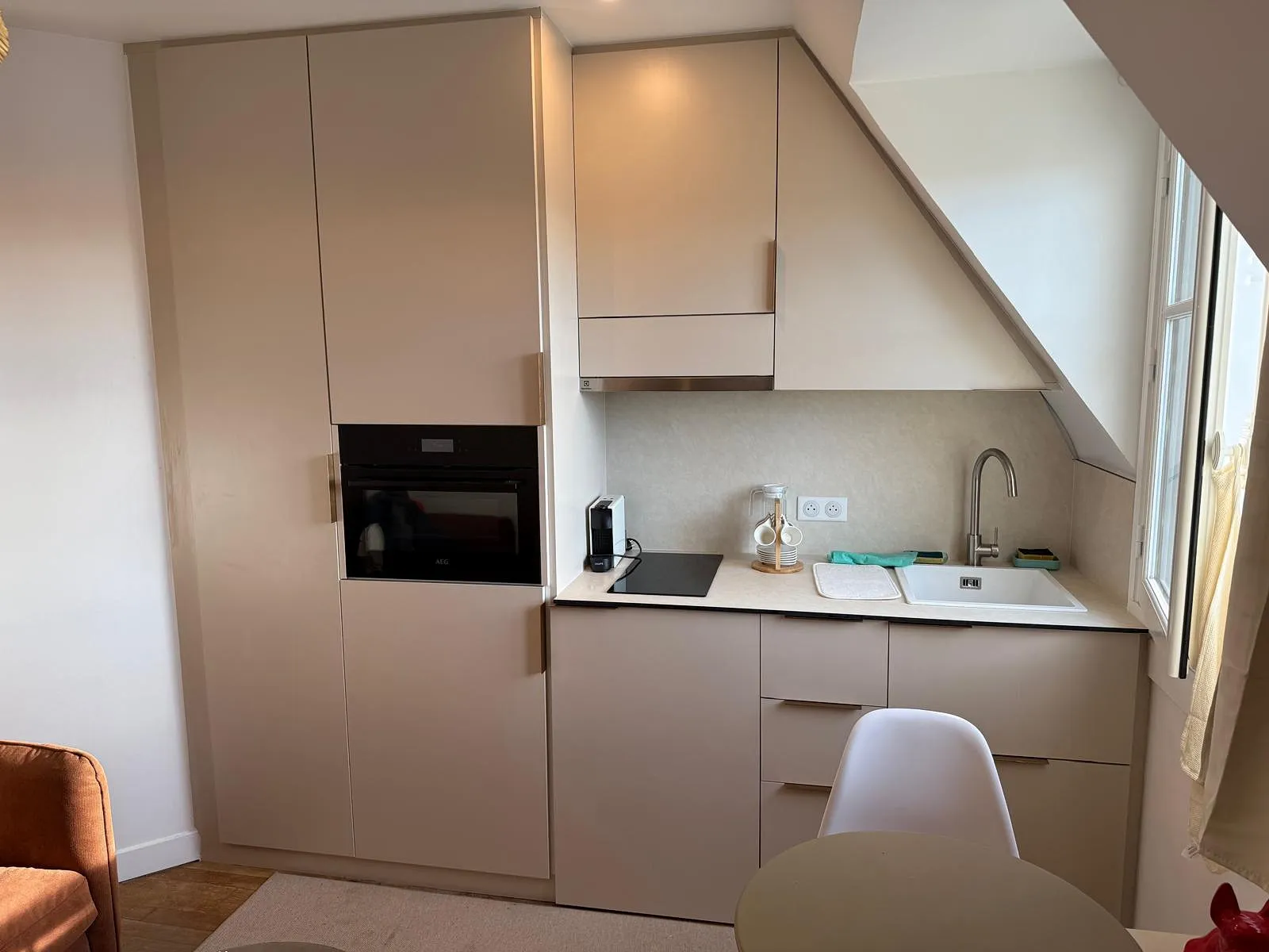 Cuisine compacte beige avec évier, plaque de cuisson, four intégré, cafetière, et fenêtre sur le côté.