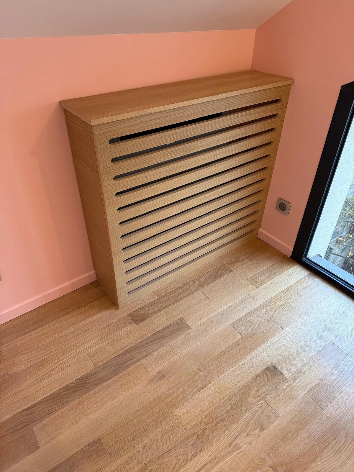 Cache radiateur en bois clair avec des lattes horizontales installé dans un coin de mur peint en rose à côté d'une fenêtre.