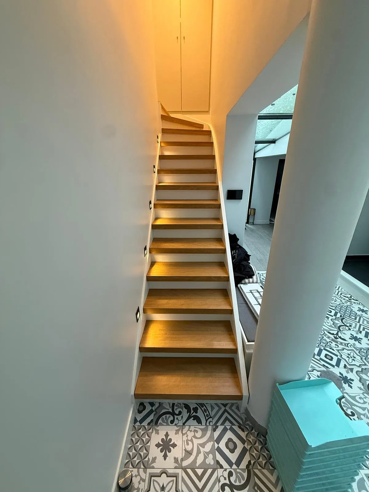 Escalier en bois éclairé incorporé dans un mur blanc avec sol carrelé à motifs géométriques et floraux.