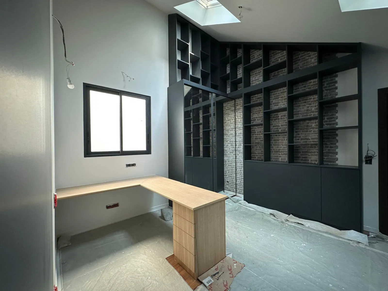 Bureau en coin avec surface en bois clair et bibliothèque noire avec étagères et miroir, sol protégé par une bâche.
