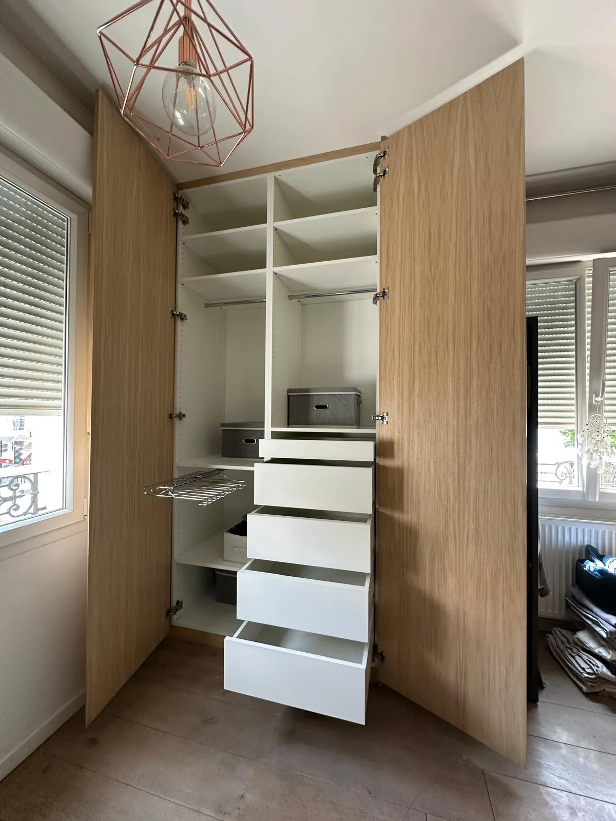 Armoire en bois clair avec portes ouvertes montrant des étagères, des tiroirs blancs ouverts, des boîtes de rangement grises et un support coulissant métallique.