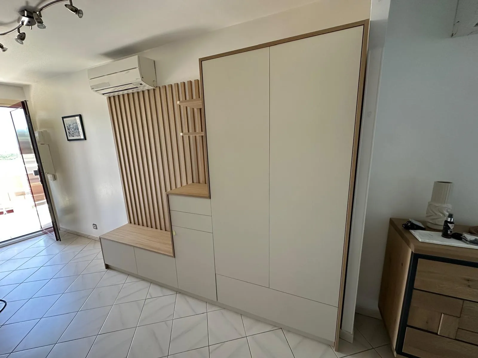 Mobilier mural moderne avec rangement beige et bois clair, comprenant un banc, des étagères, une armoire et un climatiseur au-dessus.