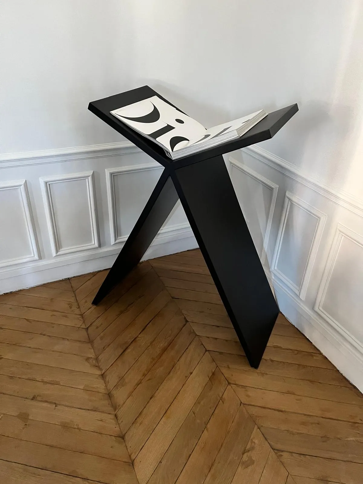 Porte-livre noir en forme de X avec un livre ouvert affichant de grandes lettres noires, placé dans un coin avec un parquet en bois et des murs blancs moulurés.